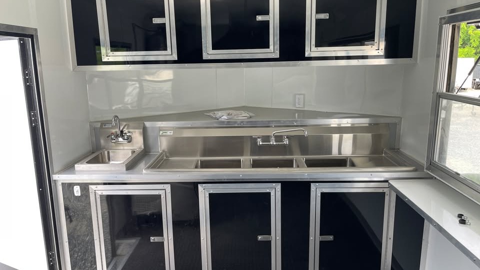 Concession trailer interior at NC Trailers with a stainless three-compartment sink, a separate handwash sink and black aluminum cabinets